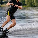 male athlete wakesurfing behind motorboat on pond, extreme summer water sports